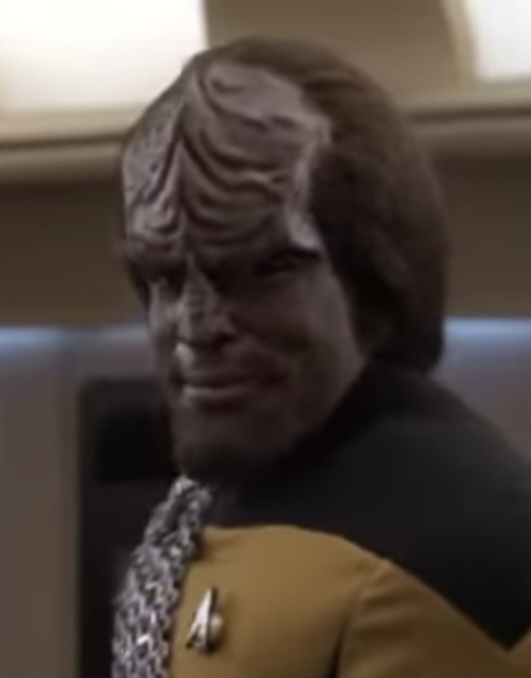 a close-up of a male Klingon character from Star Trek wearing a yellow and black uniform with a silver insignia, looking…