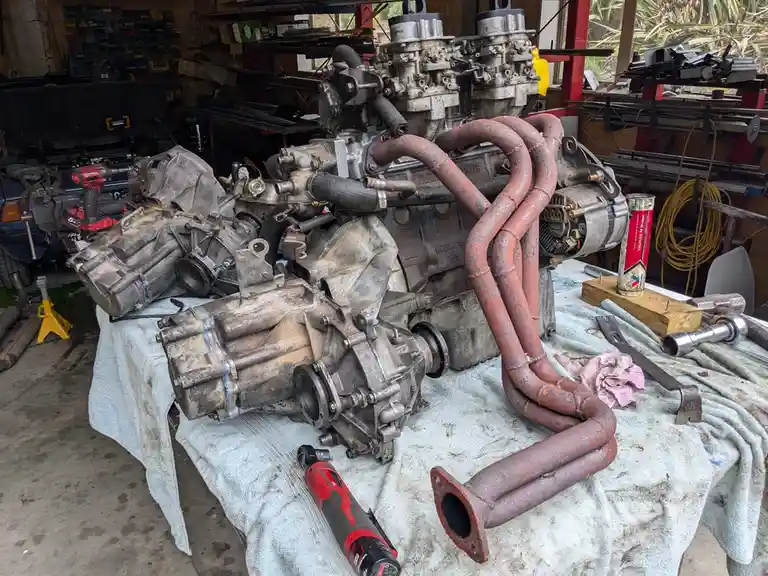 A disassembled, rusty car engine and transmission are laid out on a workbench in a cluttered garage, surrounded by tools and…
