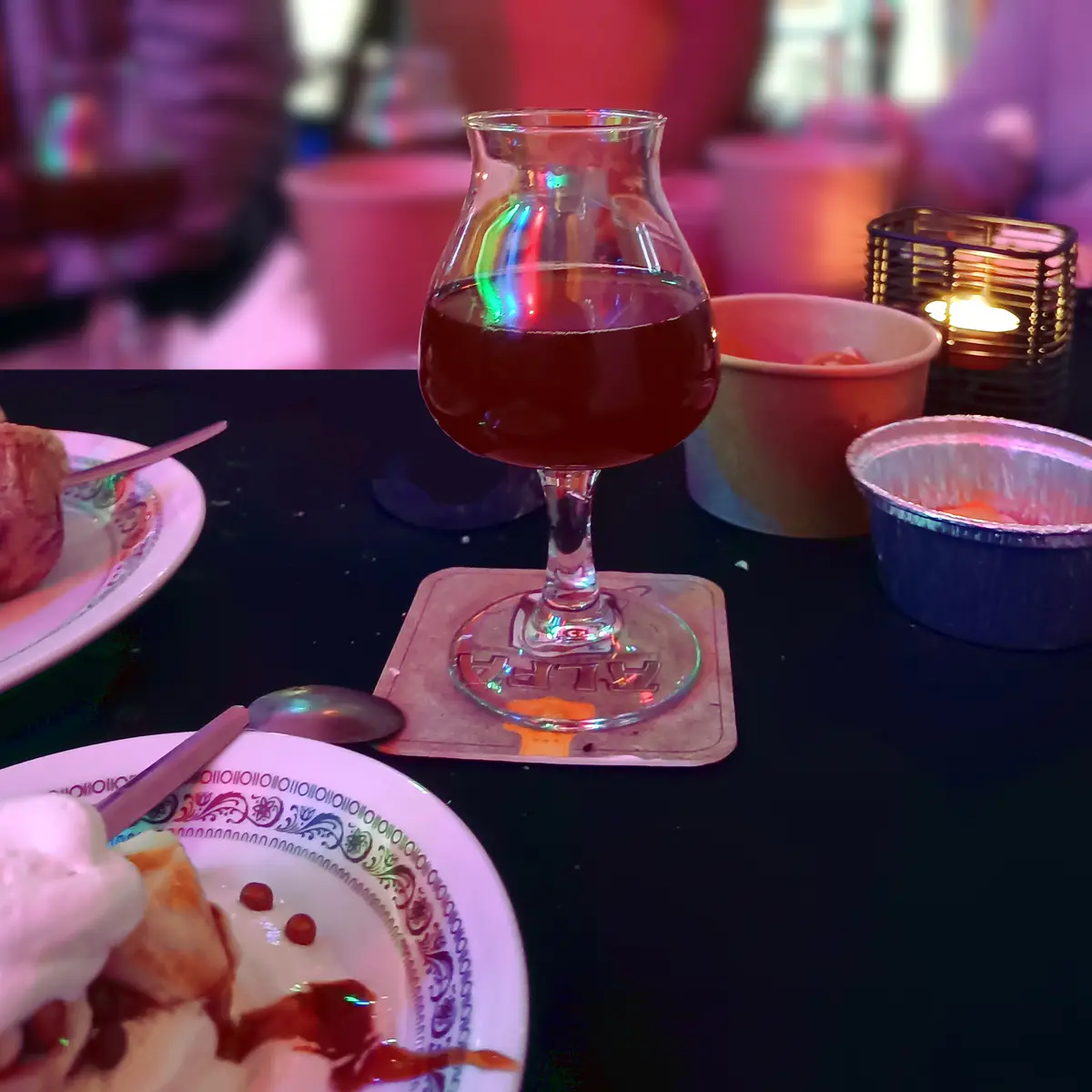 A close-up photo of a dark beverage in a stemmed glass on a coaster, surrounded by plates of dessert and small bowls, under…