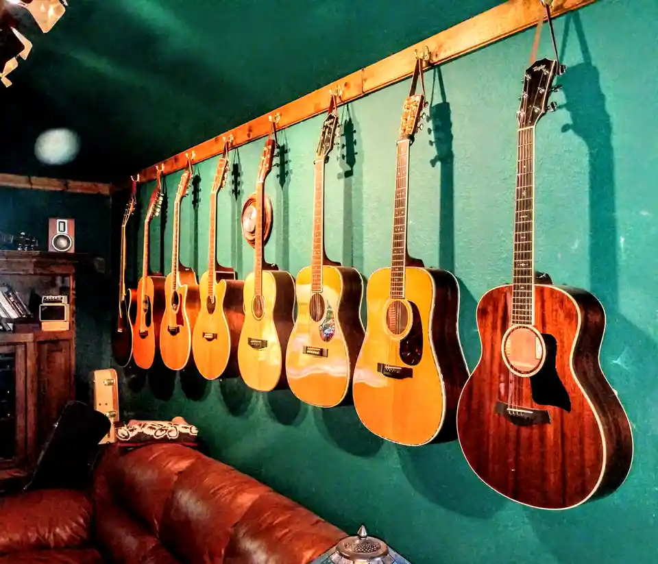 A row of acoustic guitars hangs on a wooden rack against a teal wall in a cozy room with a leather couch and vintage decor…