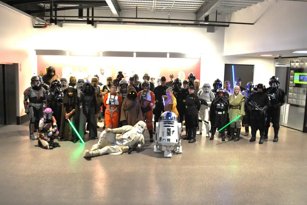 A large group of people dressed as Star Wars characters pose for a photo in an indoor hallway. In the foreground, a person…