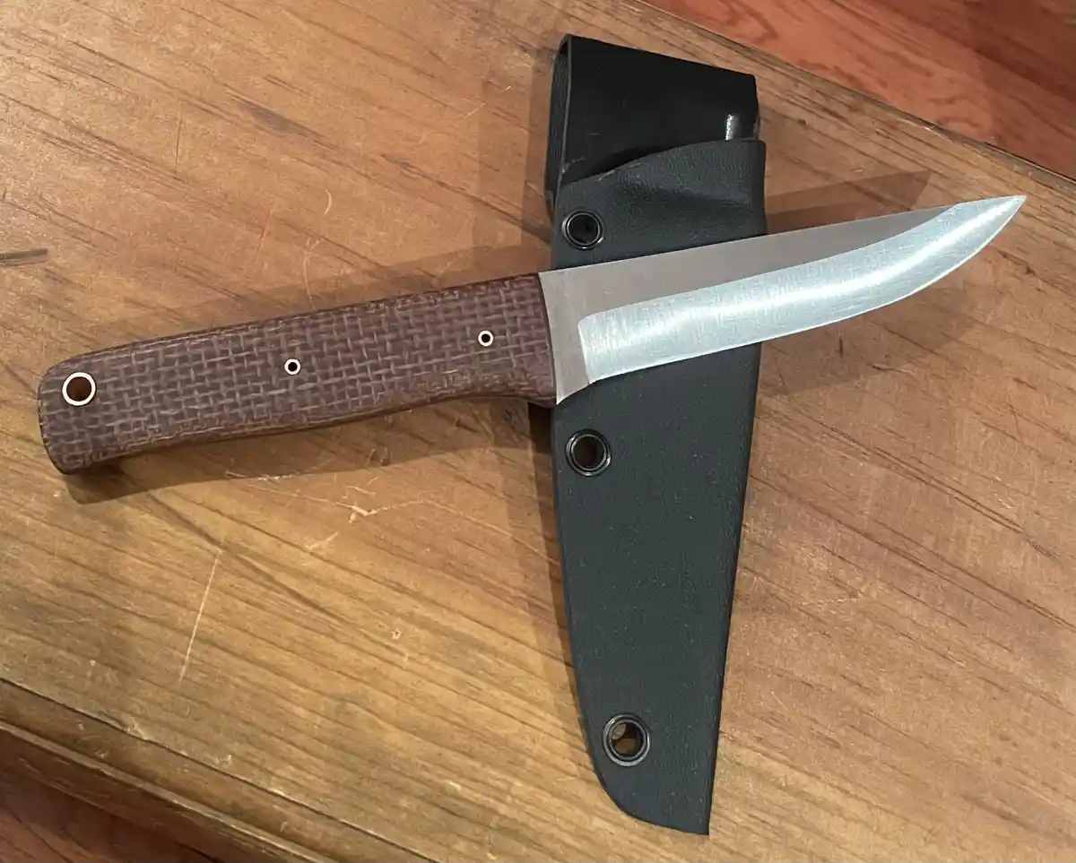 A close-up photo shows a fixed-blade knife with a brown textured handle and silver blade resting beside its black sheath on…