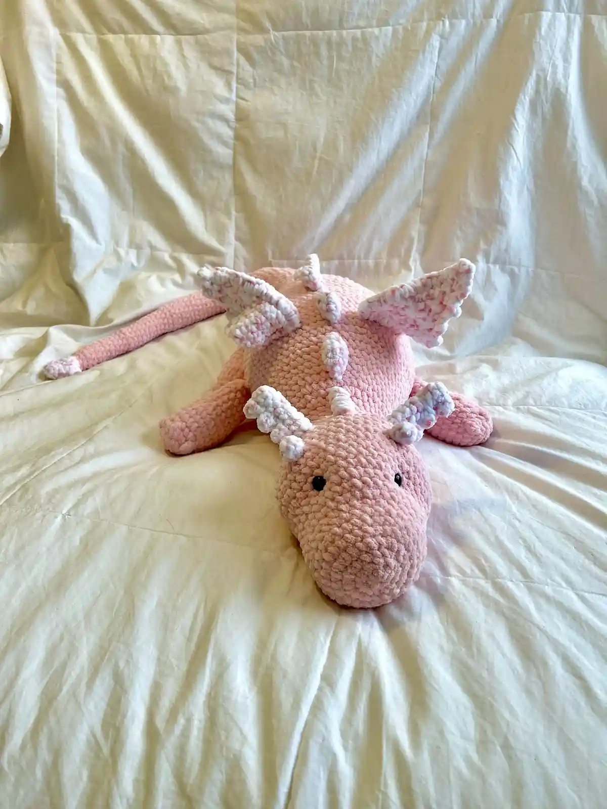 A pink, crocheted dragon plush toy with white and pink speckled wings and spikes lies on its side on a rumpled white…
