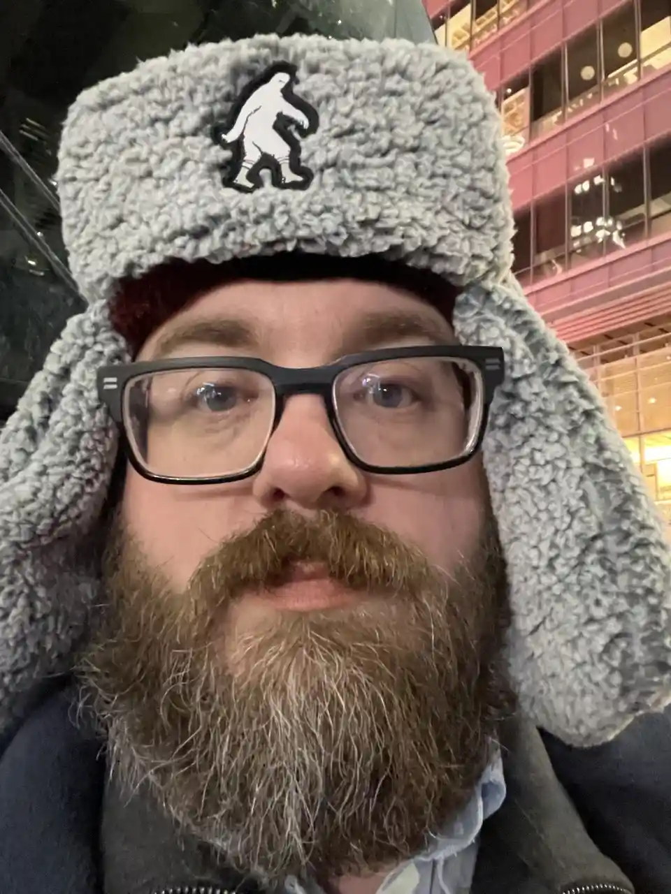 A man with a full beard and glasses wears a grey, fluffy winter hat with a black-and-white Bigfoot silhouette patch on the…