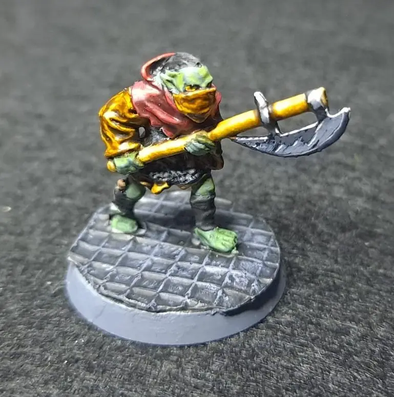 A painted miniature of a green-skinned, masked humanoid figure wearing a red hood and gold tunic, wielding a large axe with…