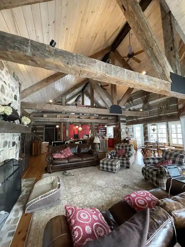 A wide-angle photo of a spacious, rustic log cabin living room with exposed wooden beams, stone fireplace, leather couches…
