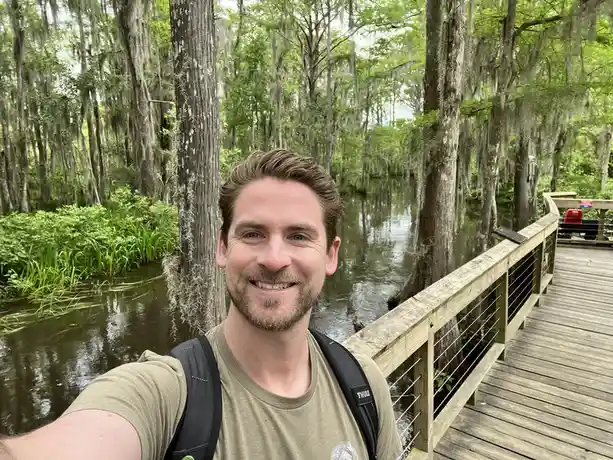A man with a backpack smiles for a selfie on a wooden boardwalk through a swamp, with tall trees draped in Spanish moss and…