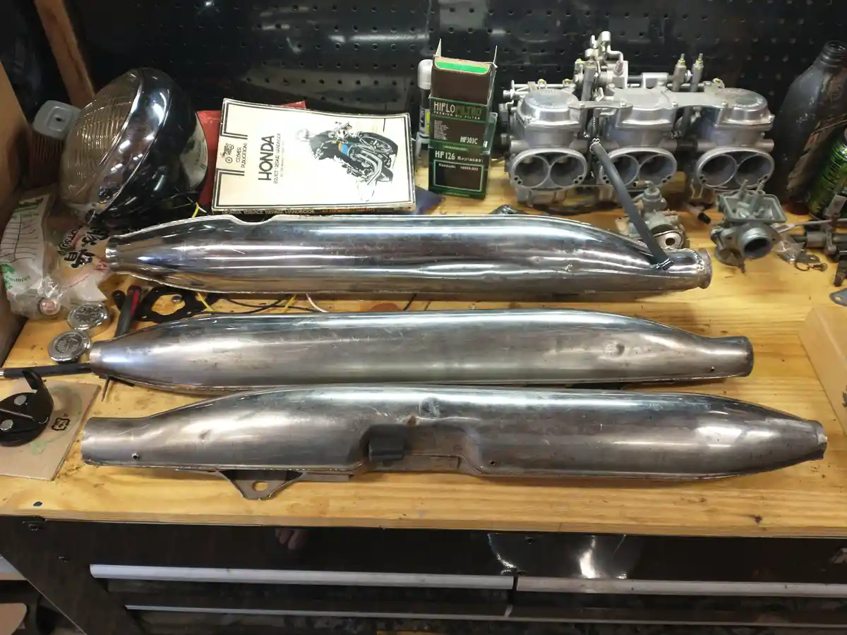 Three progressively more worn and damaged chrome motorcycle exhaust pipes are laid out on a wooden workbench, with a shiny…