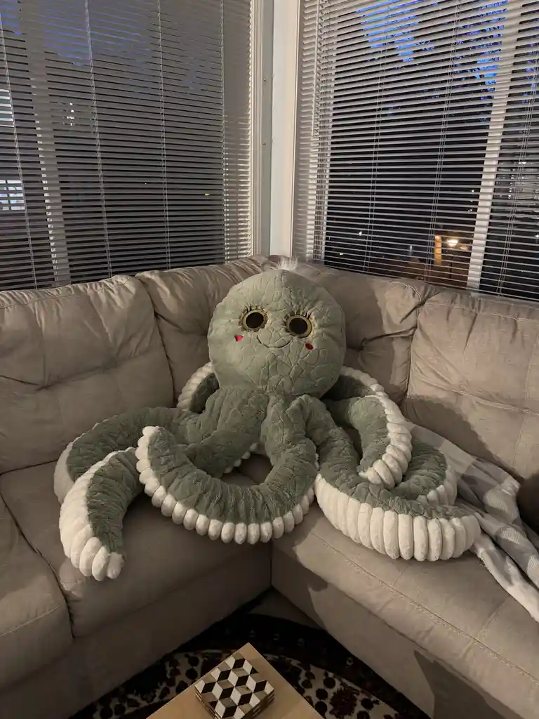 A large, light green plush octopus with big round eyes and white-tipped tentacles is sitting comfortably on a beige…