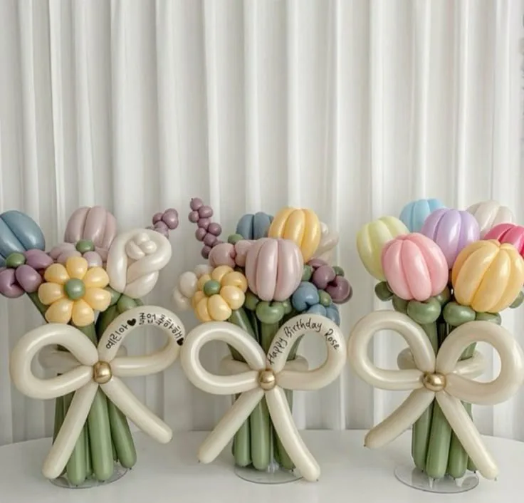 Three balloon bouquets shaped like flowers with large cream bows at the base, each bouquet has a handwritten tag. The middle…