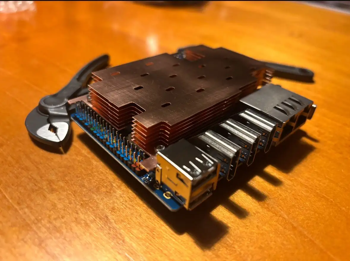 A blue circuit board with a large copper heatsink and multiple USB ports is shown on a wooden table, with a pair of pliers…