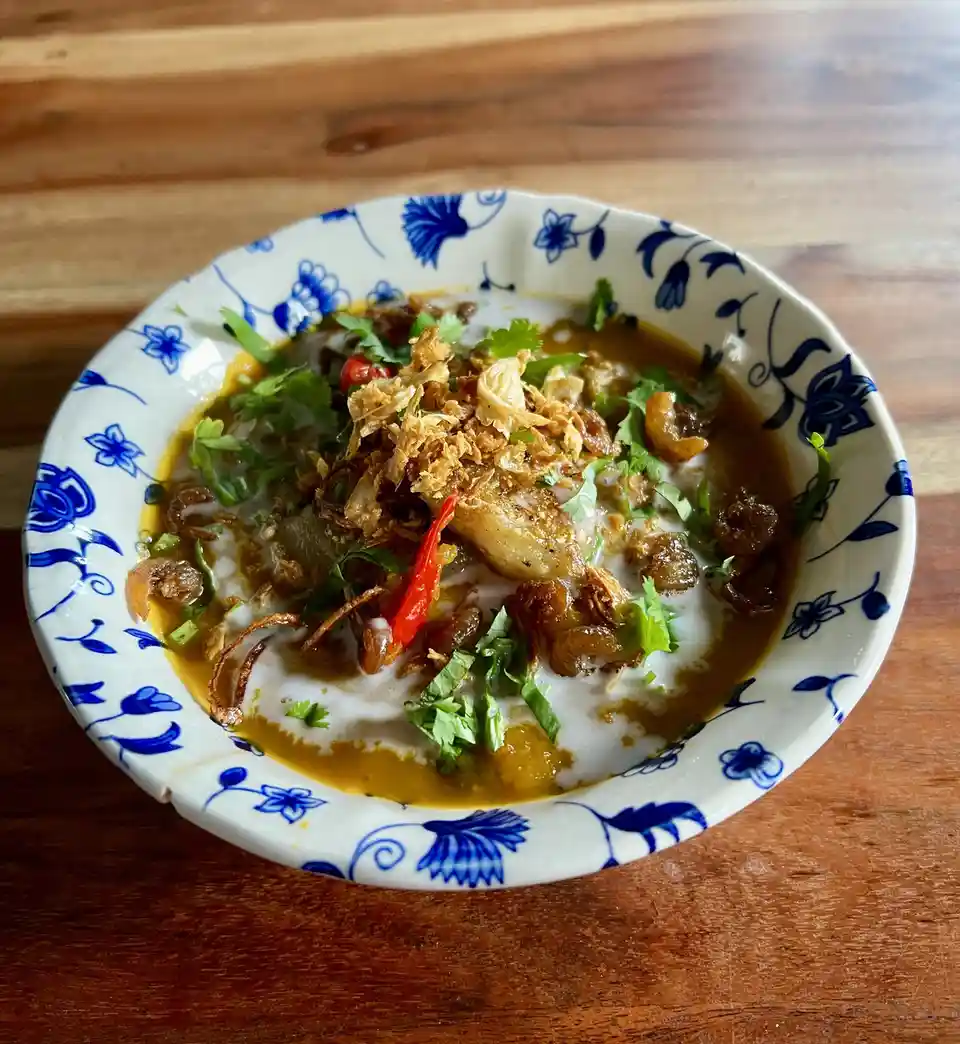 A close-up photo shows a bowl of food with a brownish-orange broth, white coconut milk, fried shallots, chopped green…