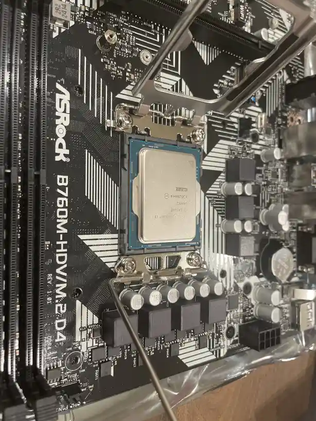 A close-up photo of an ASRock B760M-HDV/M.2 D4 motherboard with an Intel Core i3 CPU installed in its socket. A metal lever…