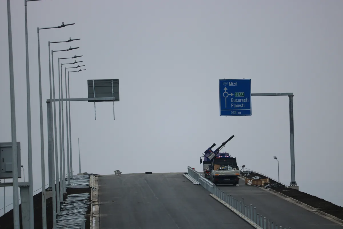 A gray, overcast day on a newly paved highway with construction materials and a truck parked on the right. A blue Romanian…