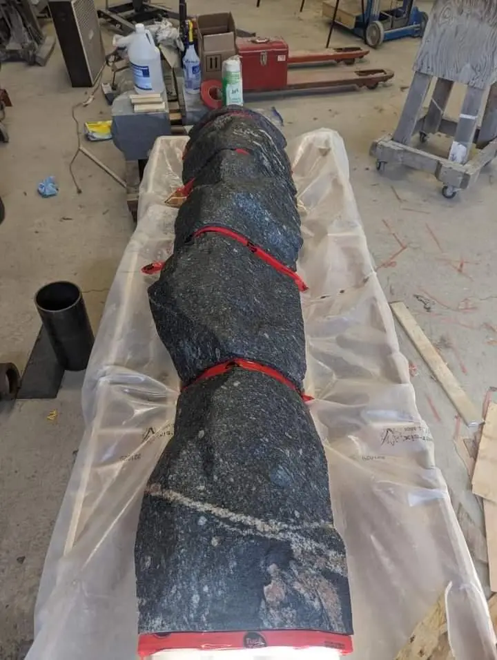 A long, dark, speckled granite slab, secured with red straps, lies horizontally on a plastic sheet in a cluttered workshop…