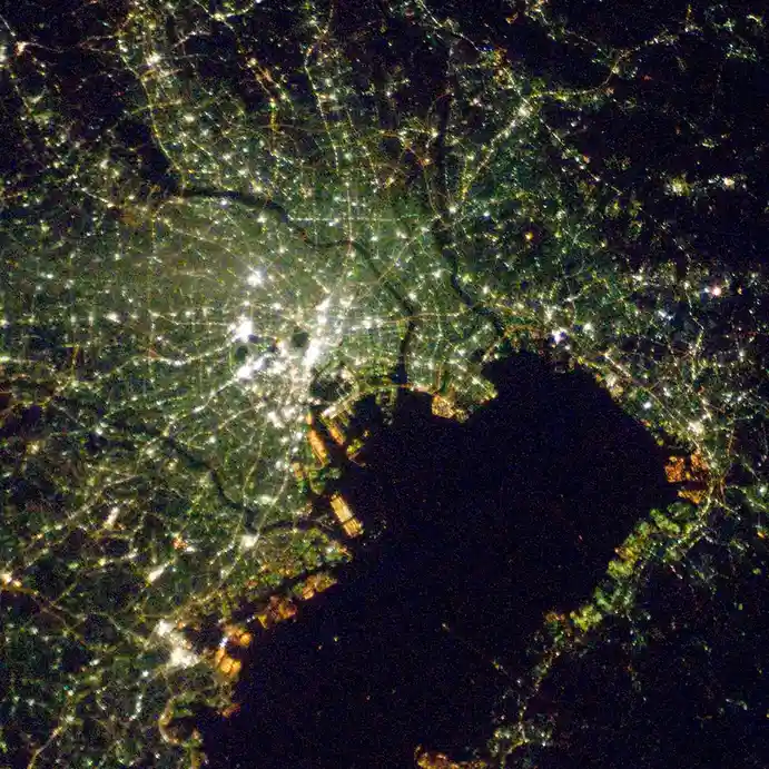 A satellite night photo shows a sprawling urban area with bright lights, where a large dark body of water is surrounded by…
