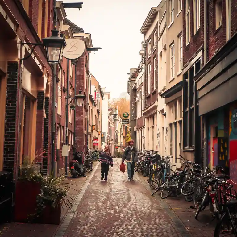 Two people walk down a narrow, cobblestoned European street lined with brick buildings, bicycles, and storefronts. The image…