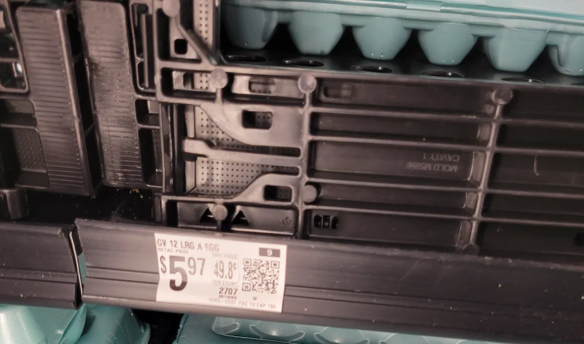 A close-up photo of an empty egg carton in a grocery store, showing the plastic mold cavities and a price tag that reads 'GV…