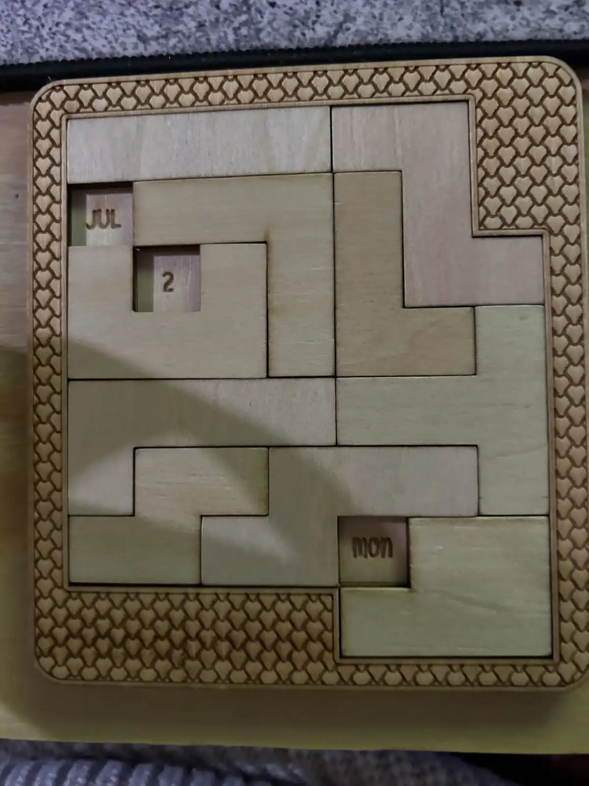 A wooden puzzle calendar with interlocking pieces, showing 'JUL' and '2' in one section and 'mon' in another, implying the…