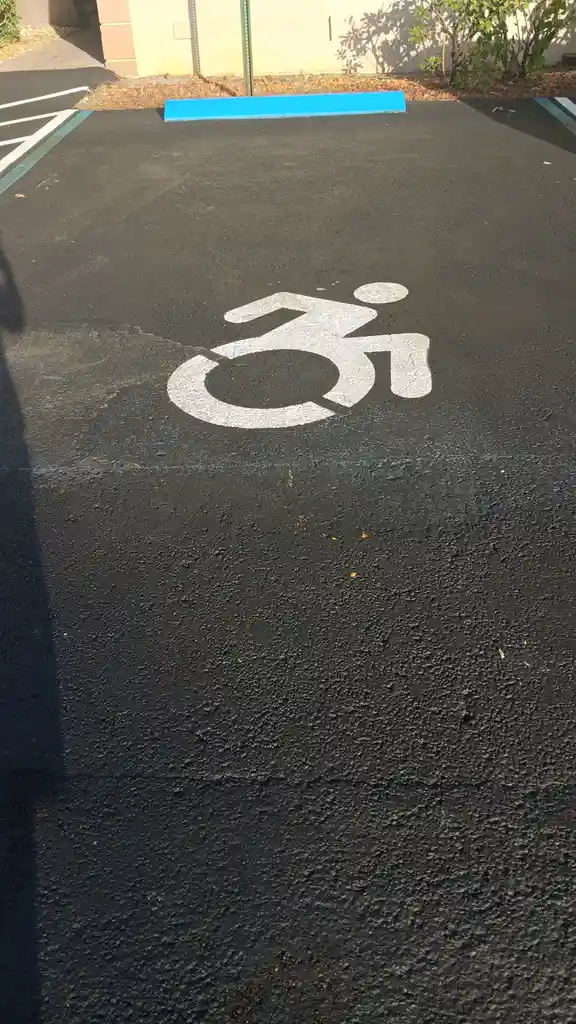 a close-up photo of a freshly painted accessible parking space with a white wheelchair symbol on black asphalt, bordered by…