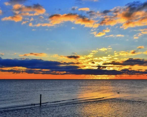 A serene sunset over the ocean with vibrant orange and blue clouds reflecting on the water, a lone surfer in the distance…