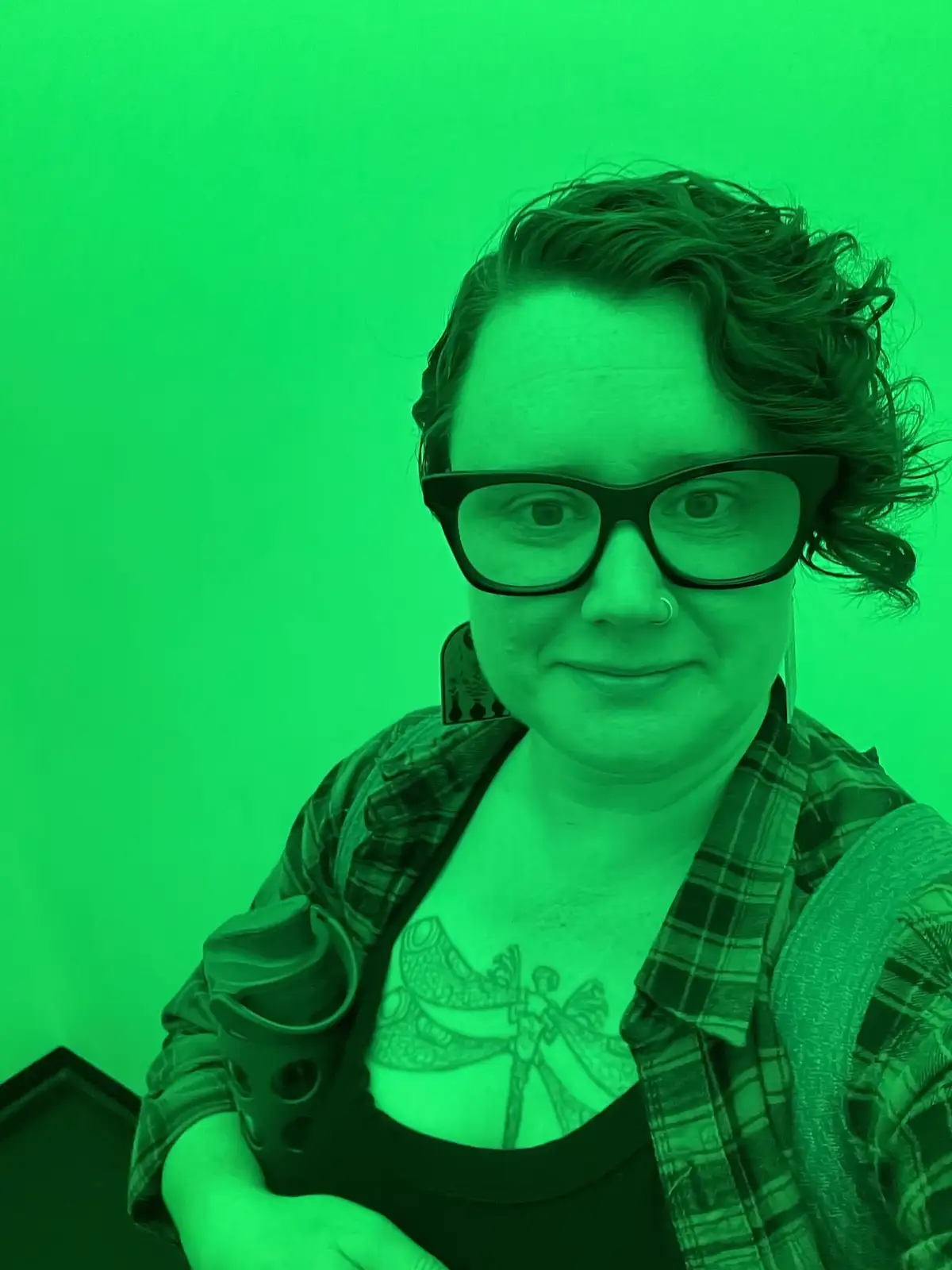 A person with short curly hair, glasses, and a nose ring smiles at the camera while wearing a plaid shirt over a black top…