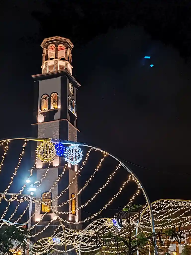 A nighttime photo of a lit-up clock tower with festive string lights and decorative globes in the foreground. The overlaid…