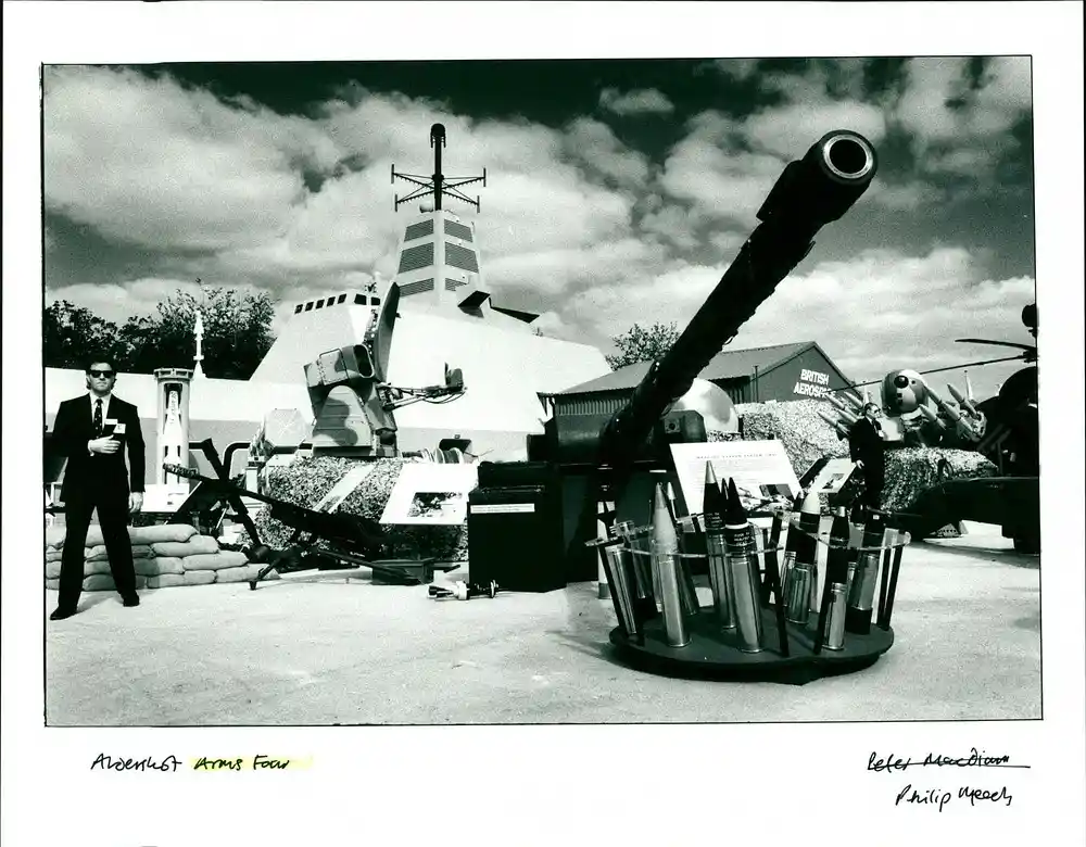 A black-and-white photograph shows a man in a suit and sunglasses standing beside a large military gun with various…