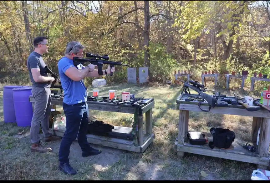 Two men are at an outdoor shooting range, one aiming a rifle with a scope while the other holds another firearm. Several…