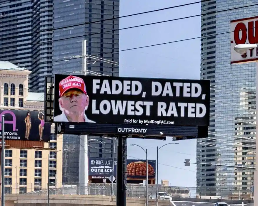 A large outdoor billboard in a city with skyscrapers shows a photo of Donald Trump wearing a red 'Make America Great Again'…