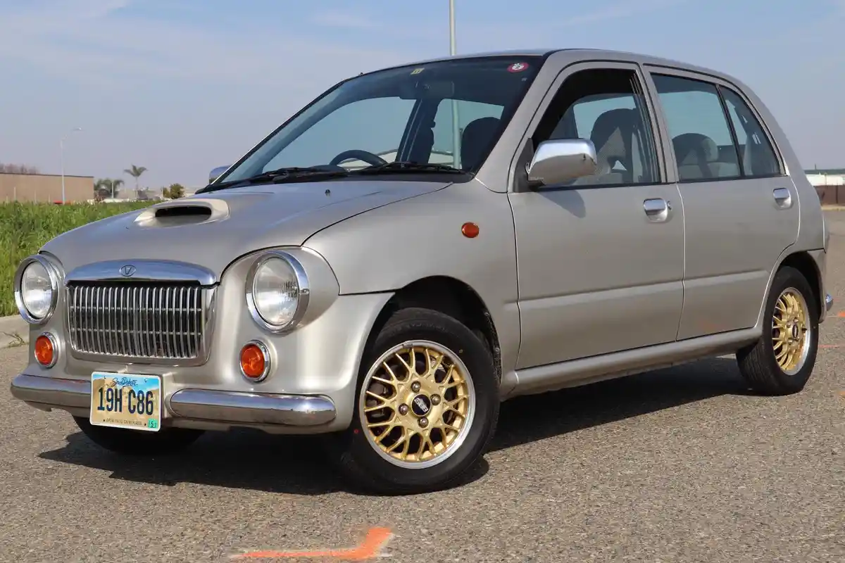 A silver compact car with a prominent hood scoop and gold multi-spoke wheels is parked outdoors. The overlaid text reads…