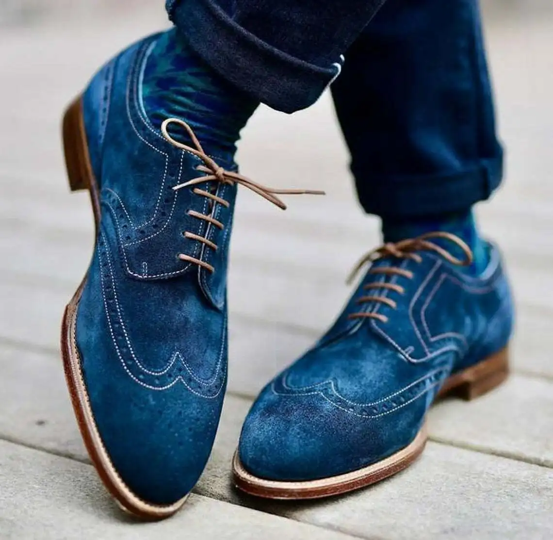 A close-up shot of a person's feet wearing stylish blue suede brogue shoes with brown laces and soles, paired with dark blue…