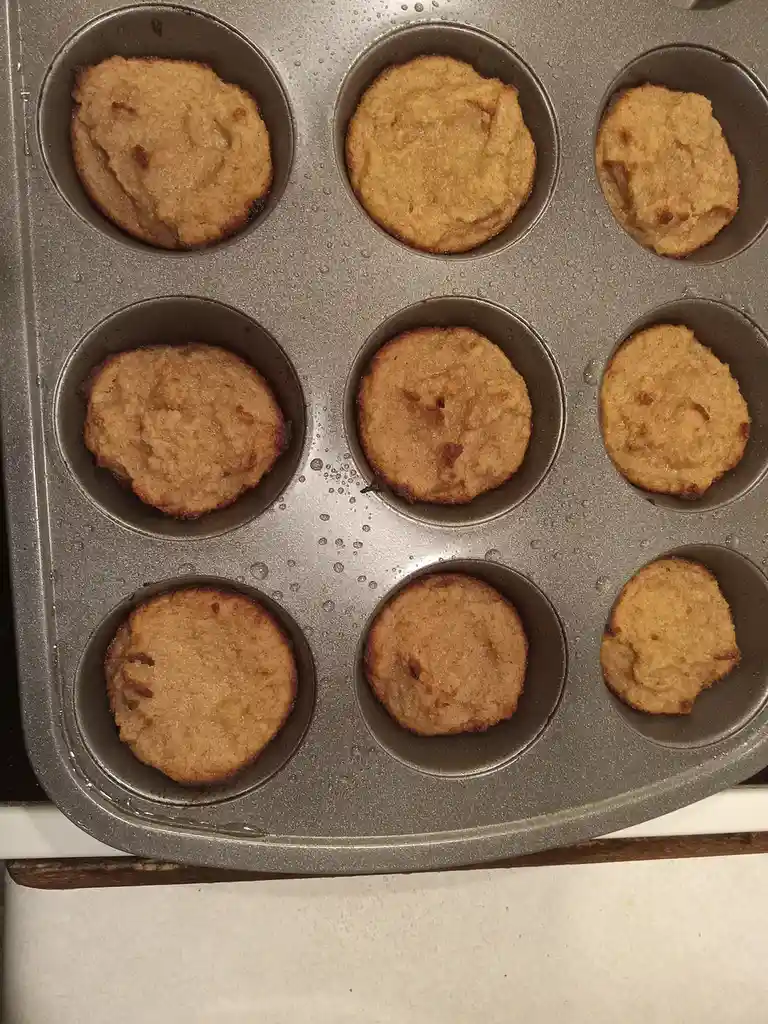 A close-up photo shows nine baked muffins in a metal muffin tin, each with a slightly uneven, golden-brown top that…