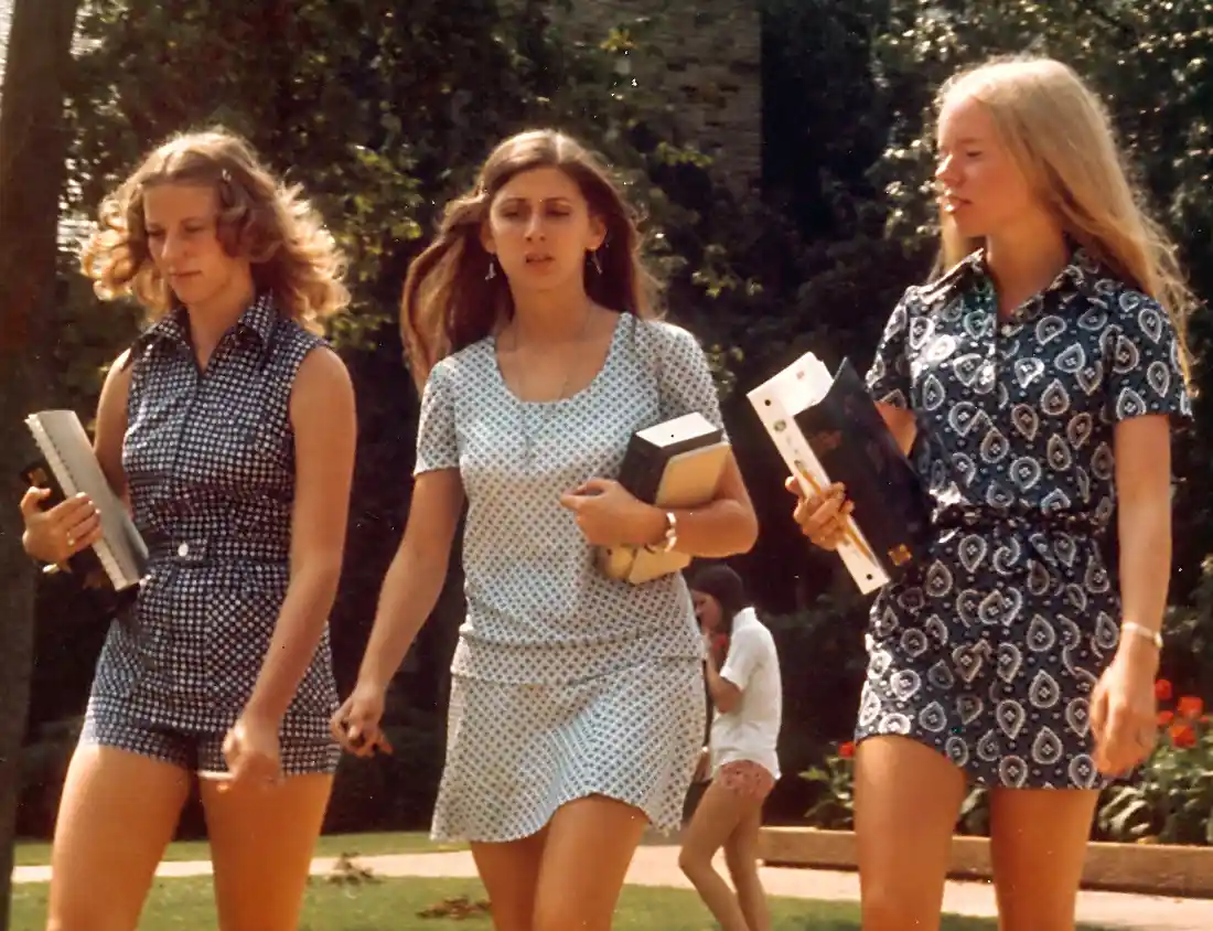 Three young women in 1970s-style patterned dresses walk outdoors carrying books and binders. The woman in the center looks…