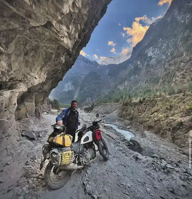 A man in a blue jacket stands beside a heavily loaded adventure motorcycle on a rocky, mountainous trail under a natural…