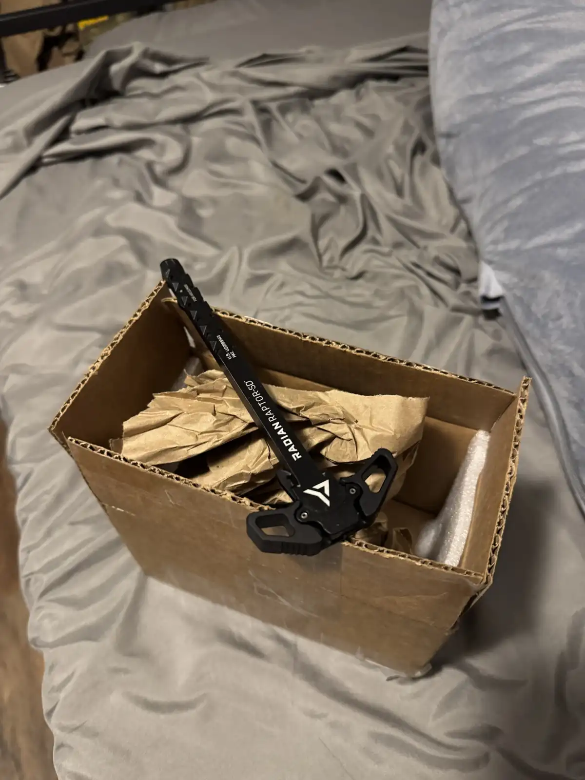 A black Radian Raptor-SD tool rests diagonally inside an open cardboard shipping box filled with crumpled brown paper and…