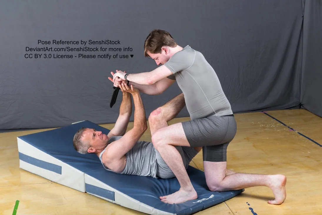 Two men are in a martial arts grappling pose on a blue mat; one man is on his back while the other is kneeling and holding…