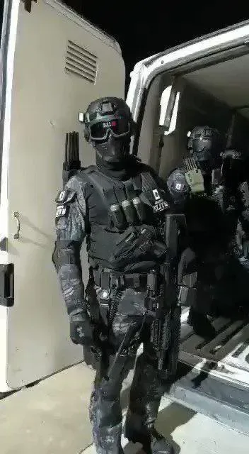 A heavily armed, fully geared soldier in tactical gear stands in the doorway of a white van at night, holding a rifle…