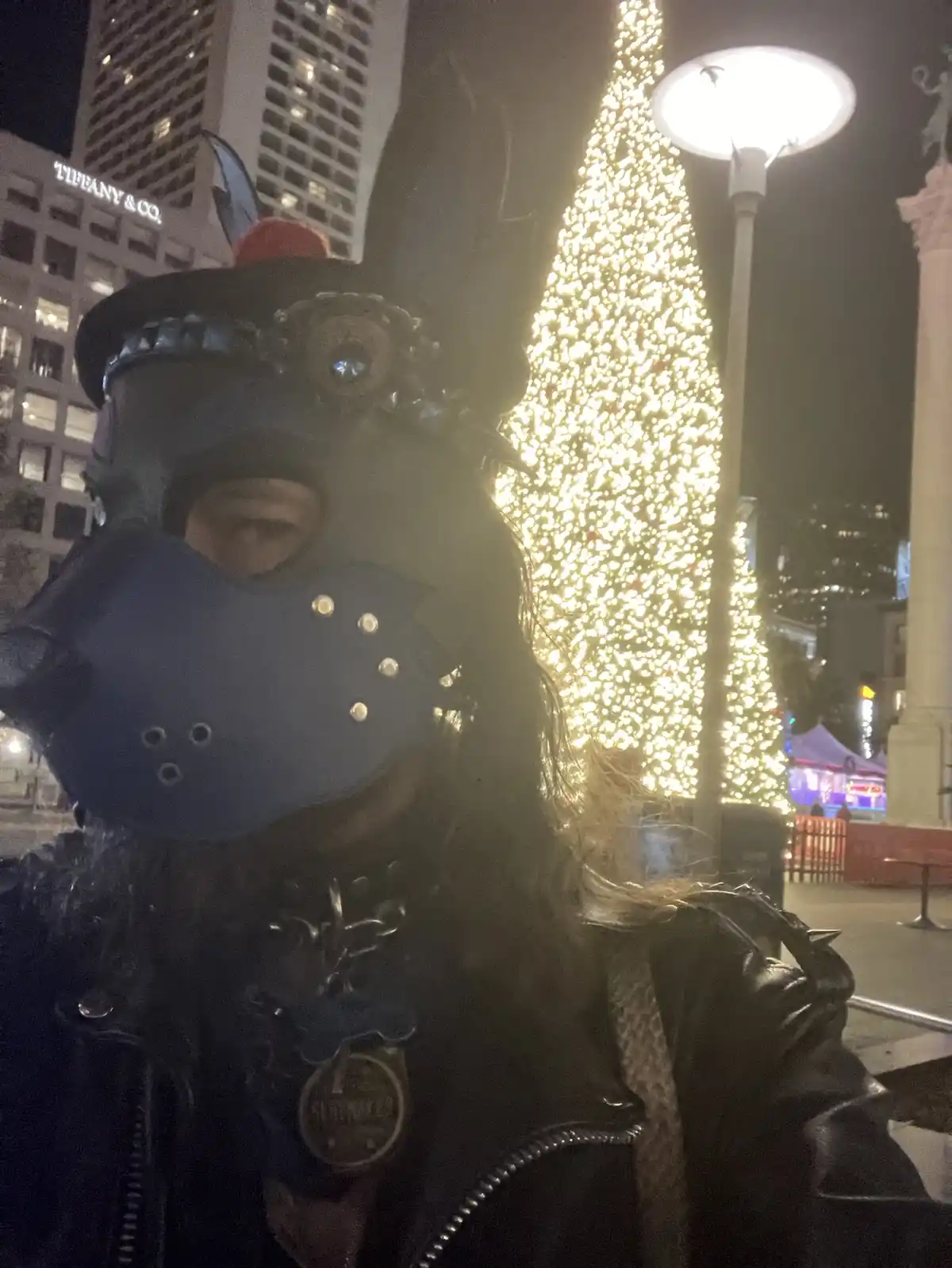 A person wearing a dark, ornate animal mask with a blue muzzle and a black hat with a red pom-pom stands in front of a…