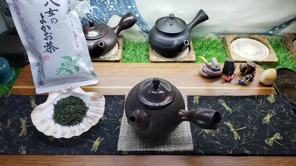 A still life of a traditional Japanese tea setup featuring a dark brown ceramic teapot on a woven mat, a white seashell…