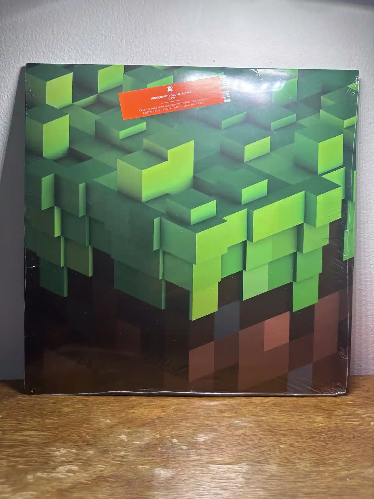 A sealed green vinyl record sleeve for 'Minecraft Volume Alpha' by C418, featuring a pixelated 3D grass block design. The…