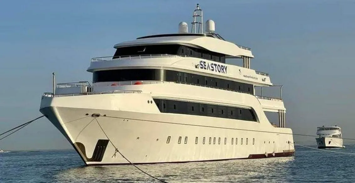 A large white luxury yacht named 'SEASTORY' is docked in calm blue water under a clear sky, with a smaller boat visible in…