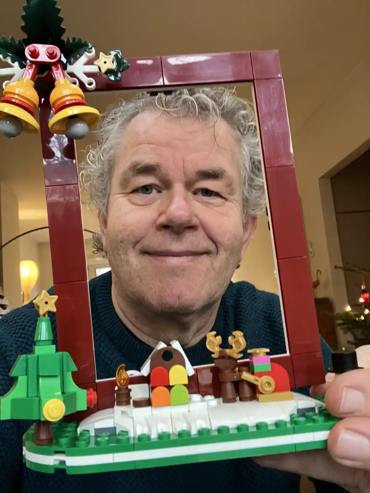 A man with curly gray hair and a blue sweater smiles while holding a red LEGO mirror frame decorated with bells, a star, and…
