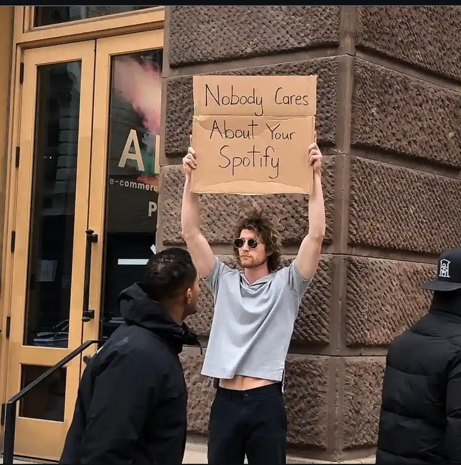 A man with curly hair and sunglasses holds up a cardboard sign that reads 'Nobody Cares About Your Spotify' while standing…