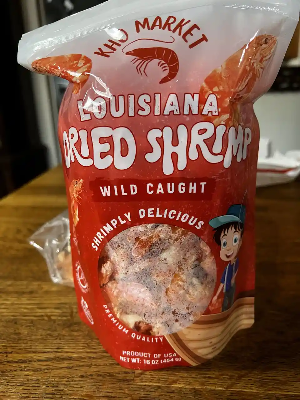 A red pouch of 'Kho Market Louisiana Dried Shrimp' sits on a wooden table, featuring cartoon shrimp, a smiling boy in a…