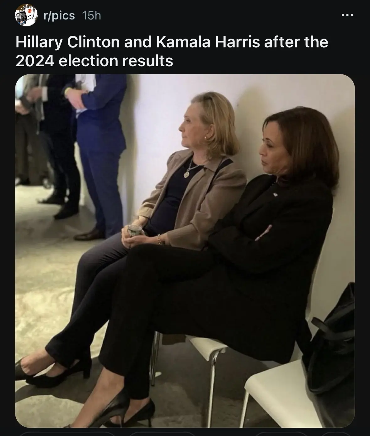 Two women, identified as Hillary Clinton and Kamala Harris, sit side-by-side on white chairs against a wall, both looking…