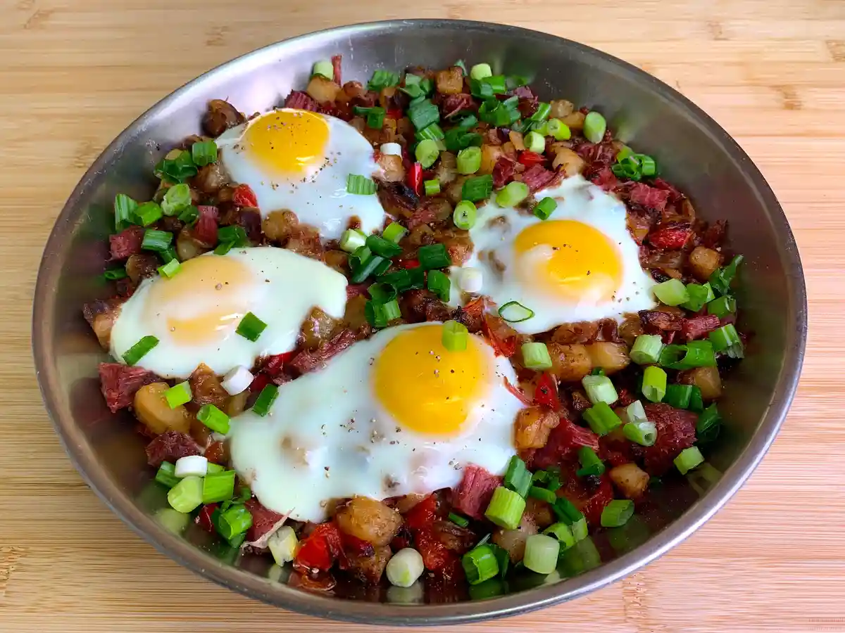 A metal bowl filled with a hearty breakfast dish featuring four sunny-side-up eggs resting on a bed of crispy potatoes…