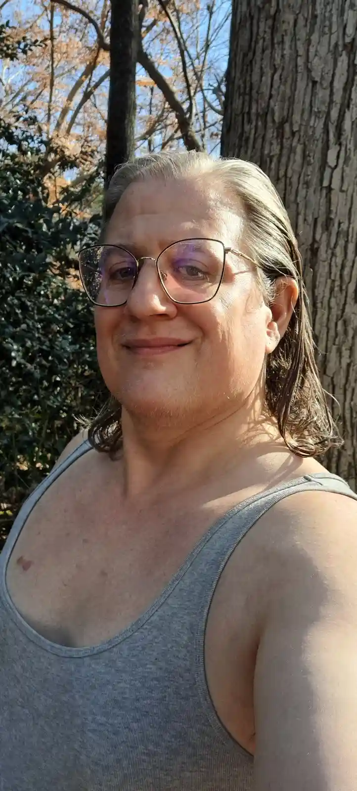 A person with long, light-brown hair and glasses smiles while wearing a gray tank top outdoors near trees. The overlaid text…
