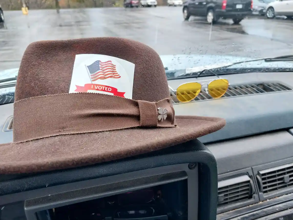 A brown felt hat with an 'I VOTED' sticker featuring an American flag is placed on a car dashboard, next to a pair of…