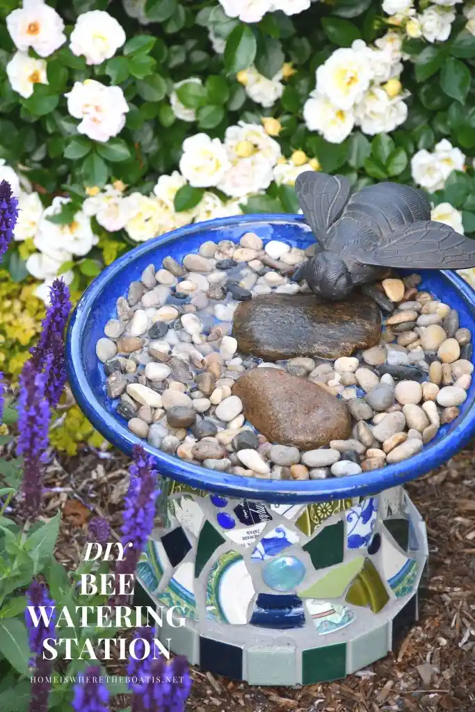 A blue ceramic birdbath filled with water and smooth pebbles, topped with a dark metal bee figurine, sits in a garden…
